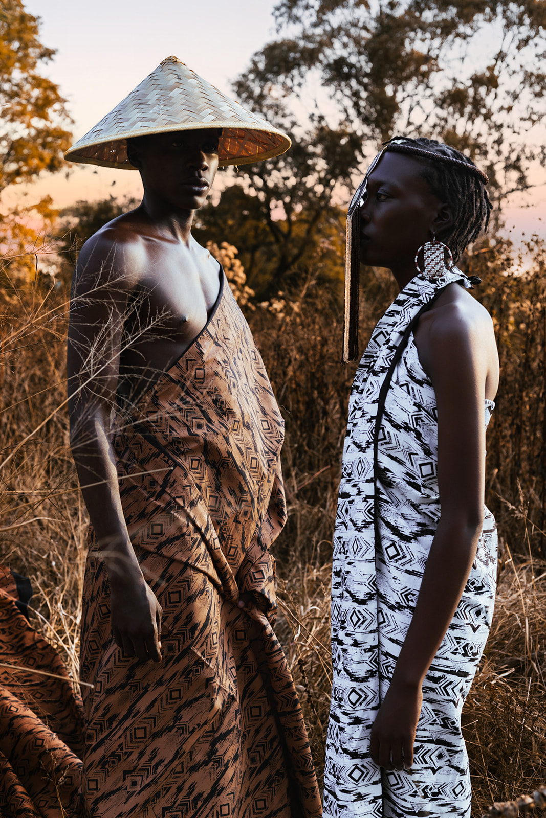 Two models in MOSI geometric wraps and hats in golden grassland at sunset, ochre and white.
