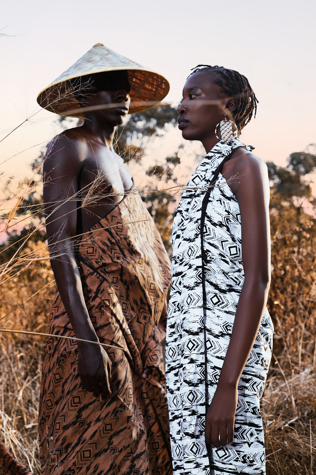Model in tan MOSI wrap and conical hat facing white-clad model, dry landscape at dusk.