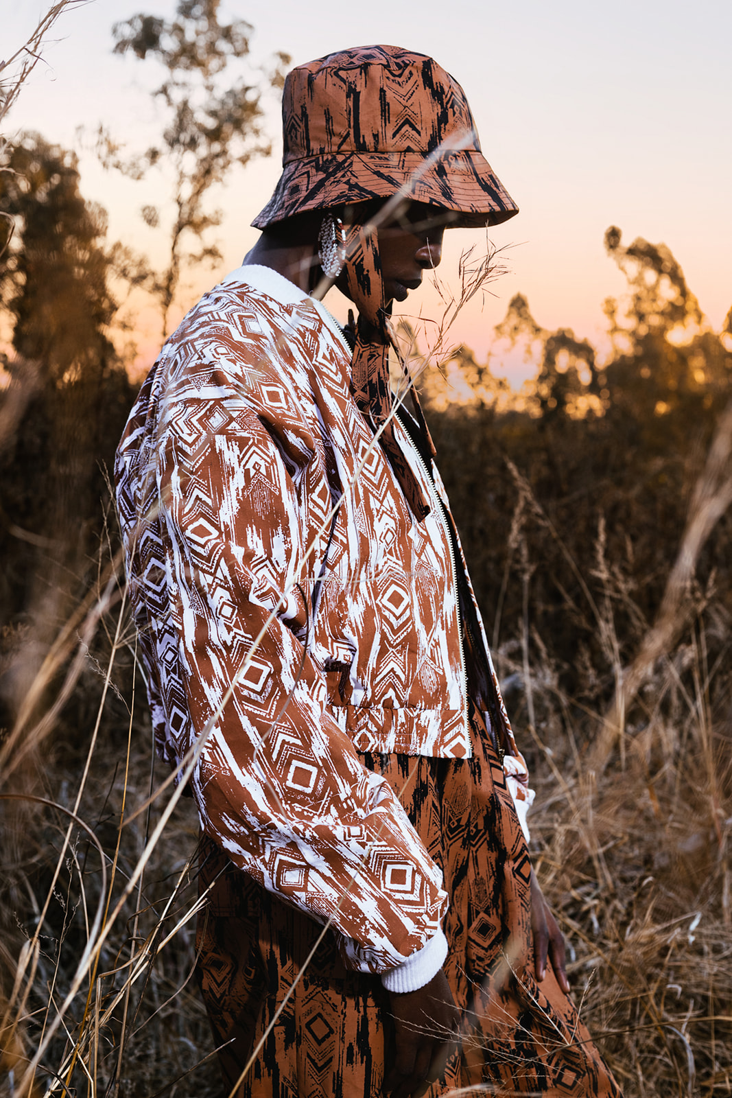Model in ochre-and-white MOSI print shirt and hat, profile pose amid tall grasses at sunset.