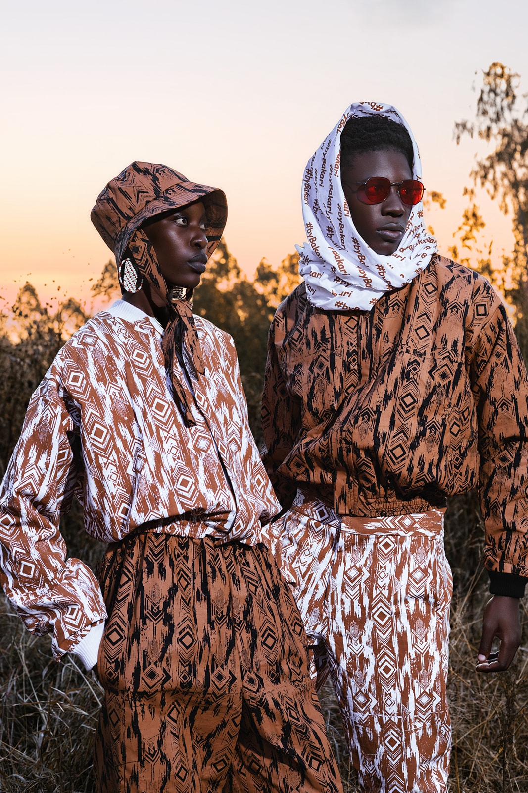 Two models in mixed MOSI print, ochre geometric and white geometric, at dusk landscape.