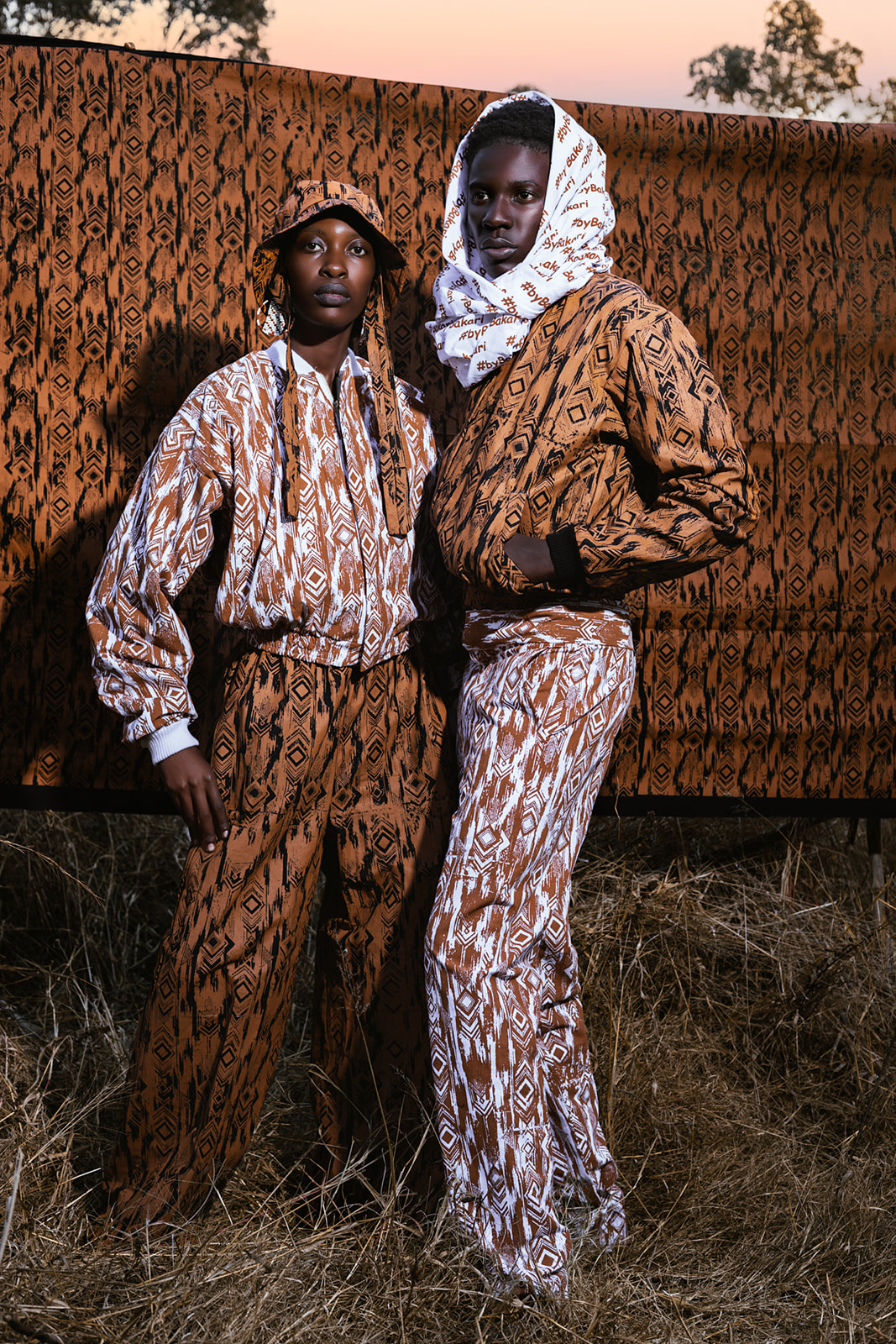 Two models in MOSI print outfits pose against weathered wooden backdrop at golden hour.