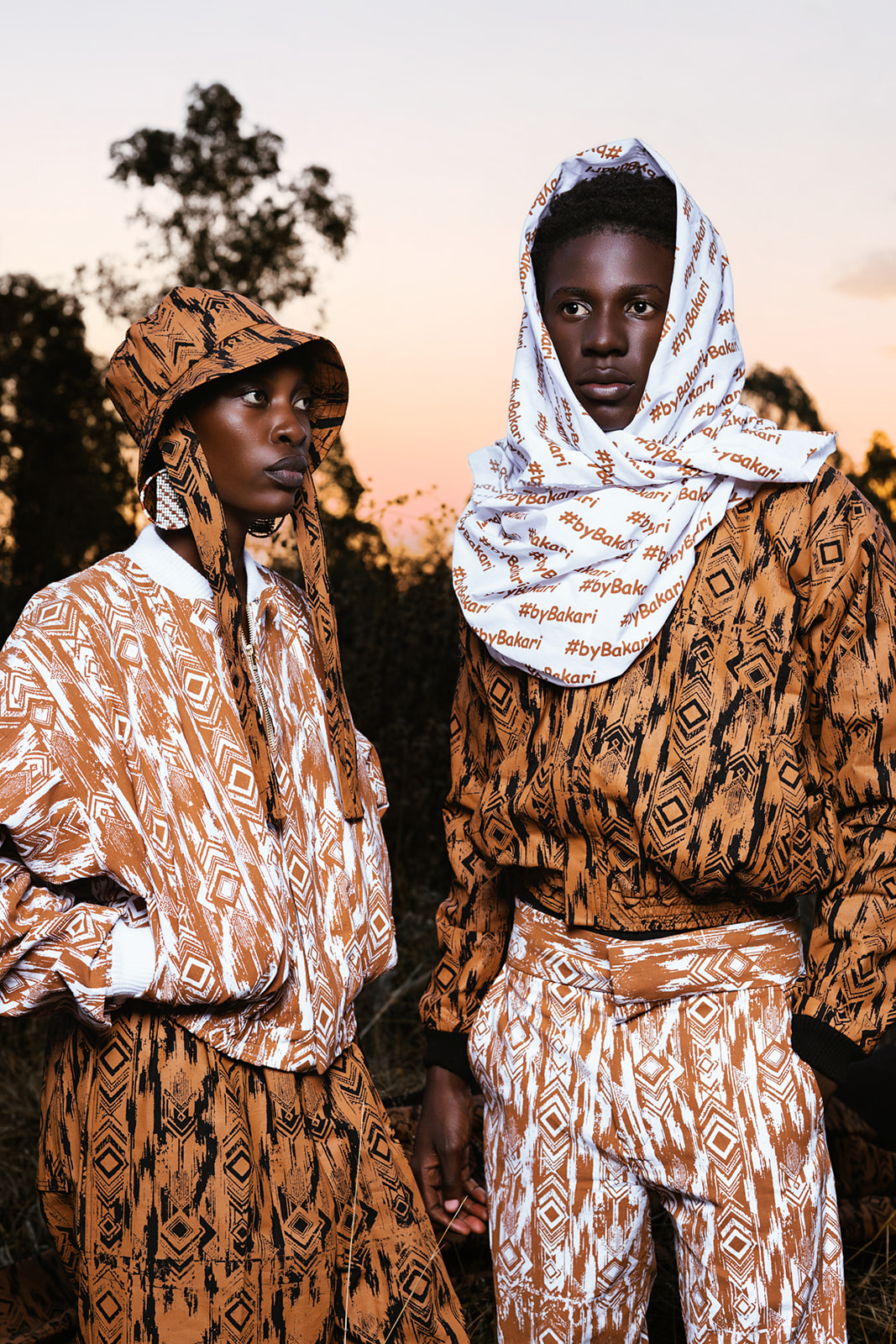 Two models in MOSI print, one white-based, one ochre, at sunset with trees silhouetted.